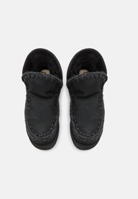 Zwarte moccasin-slippers met een zachte voering van bont, decoratieve stiksels langs de rand en een afgerond voorontwerp. Antisliprubberen zool.