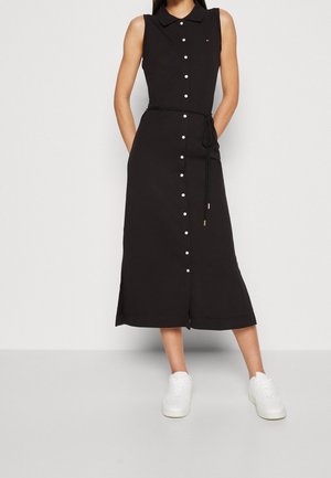 Robe midi noire sans manches à boutons avec col et ceinture à la taille, portée avec des baskets blanches.