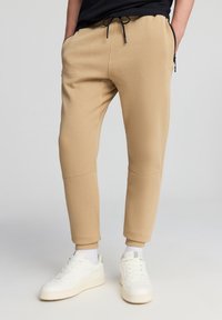 Pantaloni della tuta beige con un design affusolato, dotati di una vita con cordino, tasche laterali con zip e polsini a costine, abbinati a sneaker bianche.