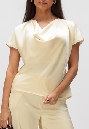 Femme portant un top court en satin crème à manches courtes avec un col drapé et un pantalon assorti, accessoirisée avec des boucles d'oreilles créoles dorées et une bague.