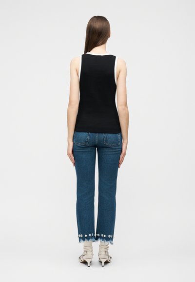 Γυναίκα που στέκεται με την πλάτη, φοράει μαύρη αμάνικη μπλούζα, μπλε τζιν με φλοράλ κρόσσια στα μπατζάκια και άσπρα σανδάλια με τακούνι.