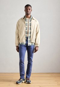 Beżowa kurtka bombowca na kratkowanej koszuli, granatowe spodnie i wielokolorowe sneakersy. Widoczne są gładkie tekstury i kontrastujące wzory.