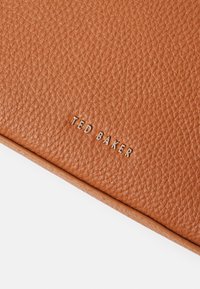 Portafoglio in pelle tan con finitura texturizzata e logo "TED BAKER" in rilievo argentato. Presenta angoli arrotondati e un bordo liscio.
