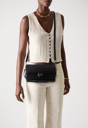 Borsa a tracolla in pelle nera con patta frontale, hardware dorato e tracolla staccabile. Giacca in tessuto testurizzato elegante e pantaloni chiari indossati.