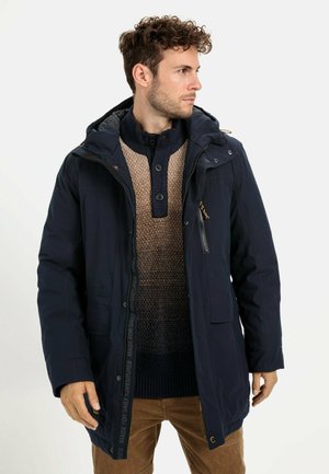 Navy blue insulated jacket with a hood, featuring a zip closure, front pockets, and a logo accent along the zipper. Textured sweater underneath.