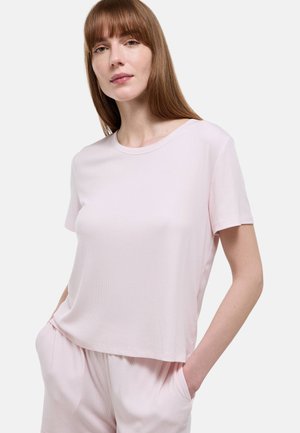 Femme aux cheveux châtain clair et raides porte un haut à manches courtes rose pâle et un short assorti, mains dans les poches, sur un fond blanc.