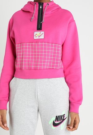Sweat à capuche court rose vif avec fermeture éclair noire, détail de poche à carreaux, et pantalon de survêtement gris avec logo Nike accentué de néon sur la jambe gauche.
