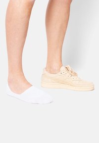 White sock and beige suede sneaker, featuring a textured upper, perforated accents, and a lace-up design; both on a light background.