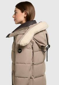 Donna di profilo che indossa un giubbotto invernale imbottito beige con cappuccio foderato in bianco e una toppa nera sul braccio.