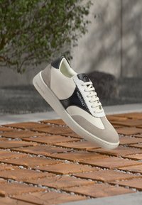 Baskets en cuir avec une empeigne blanche et beige, des accents noirs et des détails en relief. Comprend un laçage à l'avant et un logo sur le côté.