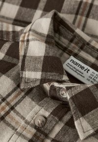 Brązowa i kremowa koszula flanelowa w kratę z kołnierzykiem, zaokrąglonym dołem i zapięciem na guziki. Tkanina ma miękki, strukturalny wygląd.
