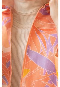 Orange jakke med blomsterprint og en lys beige ribbet rullekrave under. Har lynlåslukning og teksturerede stofdetaljer.