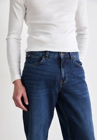 Donkerblauwe denimjeans met een rechte snit, voorzien van een klassiek vijf-pocket ontwerp en subtiele stiksels. Gecombineerd met een wit langemouwen shirt.