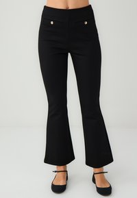 Zwarte flared broek met een gladde textuur, voorzien van twee voorzakken en gouden knoopaccenten. Enkelhoog ontwerp met een aansluitende taille.