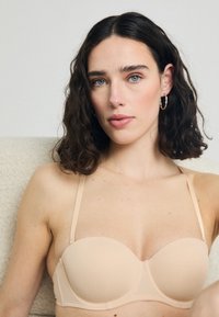Soutien-gorge sans bretelles rembourré en couleur nude, fabriqué en tissu lisse. Présente des bonnets sans coutures et des bretelles ajustables. Texture douce avec un design minimaliste.