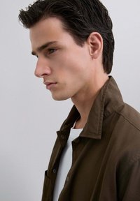 Tânăr cu păr închis, pieptănat înapoi, purtând o jachetă maro peste o cămașă albă, privind gânditor spre stânga, pe fond simplu.