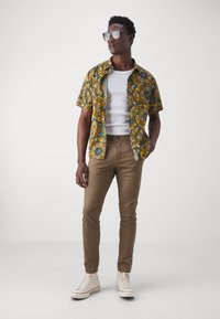 Chemise à manches courtes vert foncé avec un motif floral jaune, associée à un pantalon slim beige et des baskets montantes blanches. Lunettes de soleil noires portées.