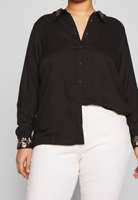 Blusa nera in tessuto testurizzato, con chiusura frontale a bottoni e accenti floreali ricamati sui polsini. Indossata con pantaloni bianchi.