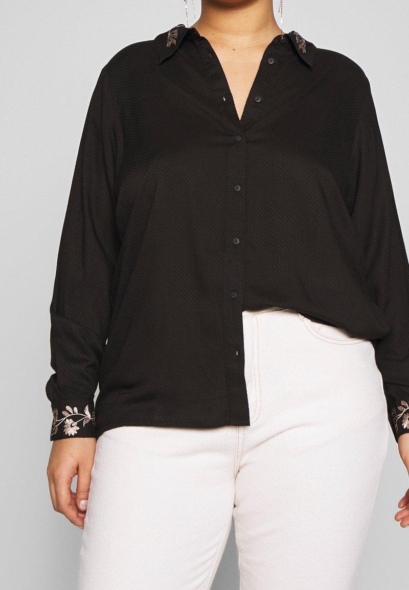 Blusa nera in tessuto testurizzato, con chiusura frontale a bottoni e accenti floreali ricamati sui polsini. Indossata con pantaloni bianchi.