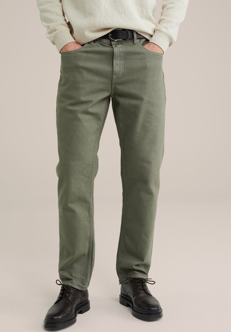 Zaļi kokvilnas bikses ar taisnu piegriezumu, ar priekšējām kabatām un pogas aizdari, kombinētas ar melniem ādas apaviem.