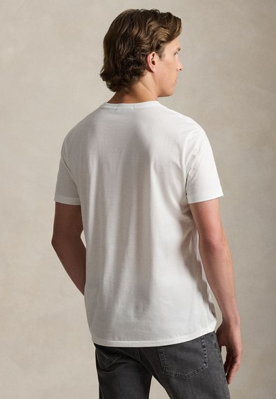 Άνδρας με κυματιστά καστανά μαλλιά φορεμένος με απλό λευκό κοντομάνικο μπλουζάκι και σκούρο τζιν, στραμμένος με την πλάτη προς το μπεζ φόντο.