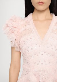 Femme portant une robe rose clair avec des manches en tulle volanté et des ornements en sequins, visible des épaules à la taille.