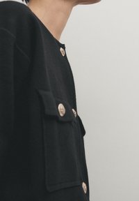 Svart skjorta med två framfickor, guldknappdetaljer och rund halsringning. Tyget verkar mjukt med en matt yta.