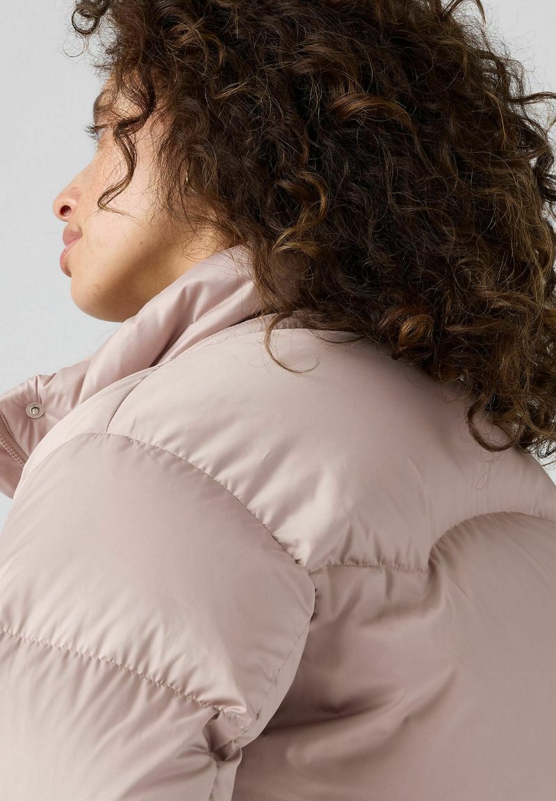 Mujer con cabello rizado oscuro de perfil a la izquierda, vistiendo una chaqueta de plumas rosa claro con un cuello alto sobre un fondo liso.