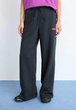 Persona vestida con pantalones deportivos negros de pierna ancha con cordón, zapatillas negras y una camisa morada, de pie sobre una alfombra azul.