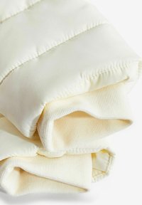 Manches d'une veste matelassée crème pâle avec des poignets côtelés tricotés rabattus sur un fond blanc.