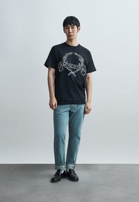 Νεαρός άνδρας στέκεται μπροστά σε απλό τοίχο, φοράει μαύρο t-shirt Moschino, ανοιχτόχρωμα μπλε τζιν με ρεβέρ, λευκές κάλτσες και μαύρα μοκασίνια.