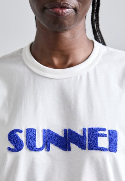 Λευκό βαμβακερό T-shirt με υφασμάτινα μπλε γράμματα που γράφουν "SUNNEI" διαγώνια στο στήθος. Κλασικός σχεδιασμός με στρογγυλή λαιμόκοψη και κοντά μανίκια.