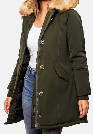 Parka femme vert foncé avec capuche bordée de fourrure, fermeture à boutons et zip, portée sur un haut blanc et un jean bleu.