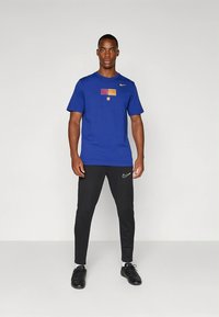 Blå Nike T-shirt med grafisk design med flag, kombineret med sorte Nike-bukser. Outfittet fuldendes med sorte træningssko.