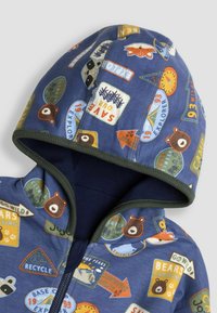 Sinine fliistöö, millel on loomade ja looduse temaatilised plaastrid. Omab roheline äärist, täispikka lukku ja kapuutsi. Pehme tekstuur ja mänguline disain.