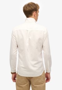 Camisa blanca de manga larga, material ligero, cuello alzado, puños con botón, diseño simple de yugo trasero, presentada con pantalones beige.