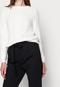 Witte geribbelde gebreide trui met open steekdetails, gecombineerd met zwarte high-waisted pantalon met een gestrikt tailleband.