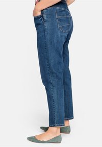 Jeans in denim blu scuro con taglio dritto, dotati di tasche posteriori e orlo sfilacciato. Abbinati a scarpe basse verdi.