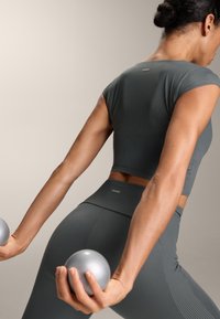 Šedý žebrovaný sportovní top a legíny s vysokým pasem. Drží stříbrné činky, ukazuje přiléhavý střih a hladkou texturu. Logo na oblečení.