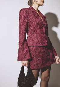 Naiselik tekstuurse tumeroosa lillemustrilise bleiseri ja sobiva miniseelikuga, hoides valge tausta ees musta helmestega käekotti.