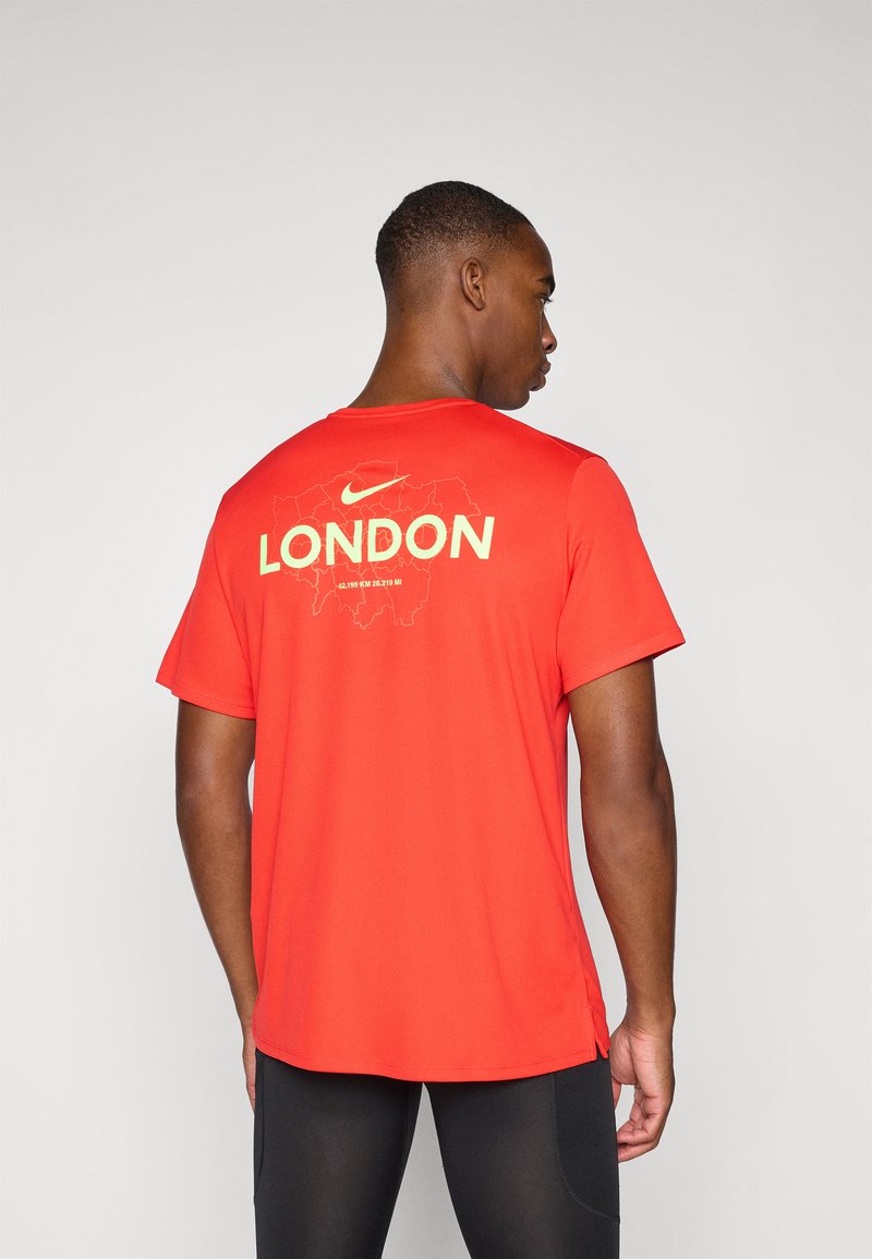 Κόκκινη μπλούζα Nike με λευκή επιγραφή "LONDON" στην πλάτη, συνοδευόμενη από περίγραμμα χάρτη της πόλης σε πιο ανοιχτό χρώμα. Έχει κοντά μανίκια και πλαϊνές σχισμές.