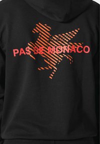 Sudadera negra con un gran gráfico naranja de un caballito de mar y el texto "PAS DE MONACO" en letras rojas en negrita.