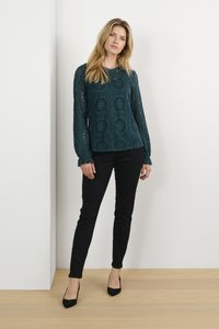 Geometrikus mintákkal díszített, hosszú ujjú, teal színű csipkefelső fekete, slim-fit nadrággal és fekete, hegyes orrú magassarkú cipővel, semleges háttér előtt.