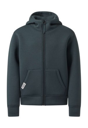 Garçon debout, main sur la nuque, portant un sweat à capuche zippé foncé, un jean bleu clair et des baskets blanches avec des rayures noires.