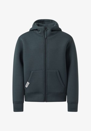 Garçon debout, main sur la nuque, portant un sweat à capuche zippé foncé, un jean bleu clair et des baskets blanches avec des rayures noires.