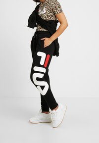 Pantalones de chándal negros con el logo de FILA en blanco y rojo, combinados con una camiseta de leopardo. Zapatillas blancas completan el look atlético.