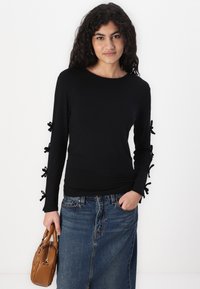 Schwarzes langärmliges Oberteil mit rundem Halsausschnitt, versehen mit dekorativen Stoffschleifen an den Bündchen. Kombiniert mit einem Jeansrock und einer braunen Handtasche.