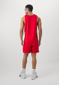 Rød treningstopp og shorts laget av mesh-materiale, med svart og hvite sideaksenter. Bært med hvite joggesko og sokker.