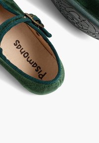 Zelená semišová slip-on obuv s kulatou špičkou, látkovým lemem a dekorativní sponou. Měkké vnitřní polstrování a texturovaná podešev jsou viditelné.