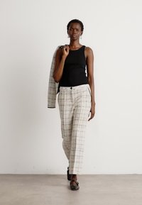 Top nero senza maniche, pantaloni crema con motivo e vestibilità rilassata, e scarpe nere. Il modello tiene una giacca abbinata su una spalla.